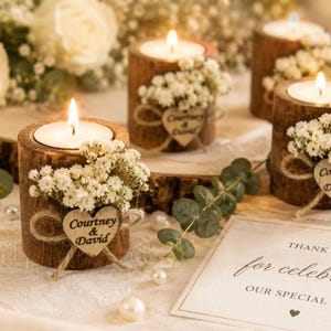 Pu&ograve; includere: Tre portacandele cilindrici in legno con corteccia esterna, ciascuno contenente un lumino acceso. Ogni portacandele &egrave; decorato con fiori bianchi di gipsofila e un piccolo cuore in legno inciso con il testo Courtney & David.