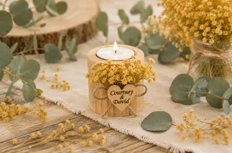Pu&ograve; includere: Un portacandele cilindrico in legno con un lumino bianco acceso. Il supporto &egrave; decorato con fiori secchi gialli e una piccola targhetta in legno a forma di cuore legata con dello spago, incisa con il testo Courtney & David.