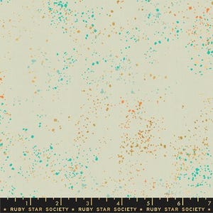 May include: Cream-colored fabric with a speckled pattern. The fabric is covered in small, irregular dots of teal, orange, and gold. The bottom of the fabric has a black ruler with the text "RUBY STAR SOCIETY".