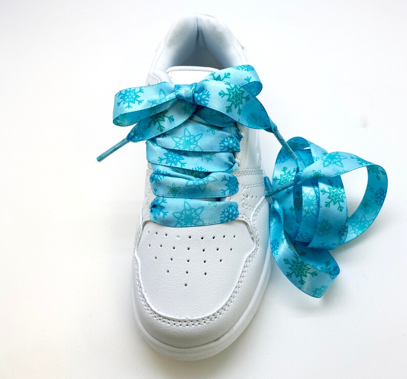 May include: White sneakers with blue snowflake patterned shoelaces tied in a bow. The shoelaces are long enough to wrap around the ankle.