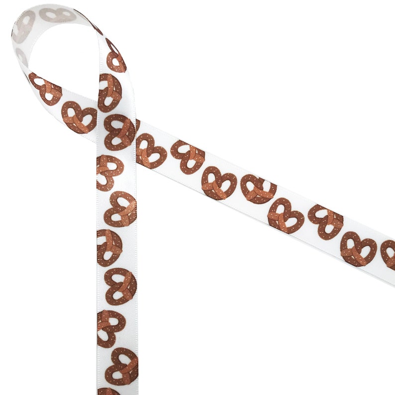 May include: White ribbon featuring a repeating pattern of brown pretzel illustrations. The pretzels have a textured appearance and are evenly spaced along the ribbon. The ribbon is folded over itself in the image.