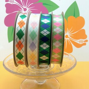Puede incluir: Cuatro carretes de cinta con un patr&oacute;n de diamante. Las cintas son verde, rosa, azul y amarilla con diamantes blancos. Las cintas est&aacute;n sobre un pedestal de vidrio sobre un fondo amarillo.