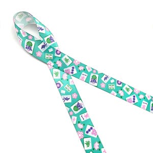 May include: A teal ribbon featuring a repeating pattern of Mahjong tiles, pink flowers, and other symbols. The ribbon is made of a smooth material and is ideal for crafting or gift wrapping. The design includes various colors, including white, green, pink, and purple.