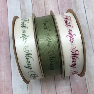May include: Three spools of satin ribbon in cream, olive green, and off-white. Each ribbon is printed with the words "Noël Merry Christmas" in a decorative font. The spools are made of cardboard.