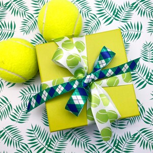 Puede incluir: Una caja de regalo verde con una cinta a cuadros blanco y azul y una cinta blanca con dise&ntilde;os de pelotas de tenis. La caja est&aacute; sobre un fondo blanco con dise&ntilde;os de hojas de palmera verdes.