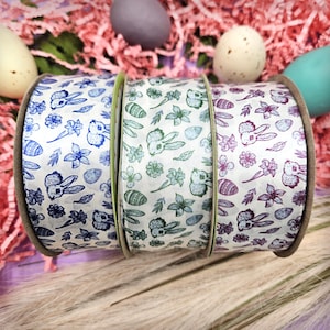May include: Three spools of decorative ribbon with Easter-themed patterns. Each ribbon features a white background with repeating designs of bunnies, flowers, and eggs in blue, green, and purple. The spools are displayed against a backdrop of Easter decorations.