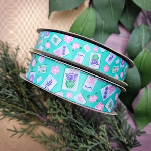 May include: Two spools of turquoise ribbon with a repeating pattern of pink flowers and Mahjong tiles. The ribbon is on a cardboard spool and is surrounded by green foliage. The Mahjong tiles have various symbols and characters.