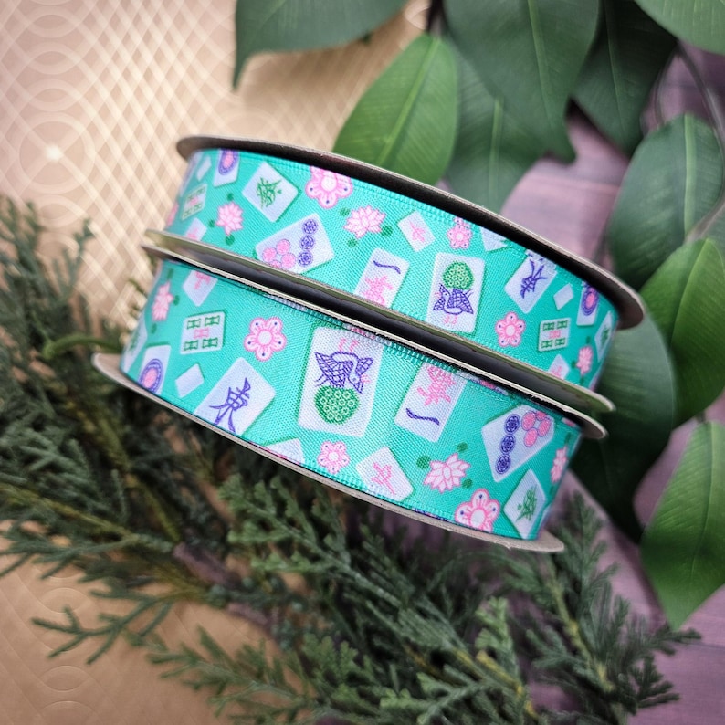 May include: Two spools of turquoise ribbon with a repeating pattern of pink flowers and Mahjong tiles. The ribbon is on a cardboard spool and is surrounded by green foliage. The Mahjong tiles have various symbols and characters.