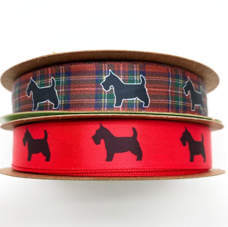 May include: Two spools of ribbon. The top spool features a plaid pattern in shades of brown, green, and blue, with black dog silhouettes. The bottom spool is red with black dog silhouettes. Both spools are on cardboard.