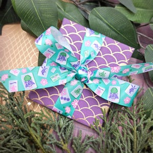 May include: A wrapped gift box with a purple and gold scale pattern, topped with a teal ribbon featuring Mahjong tiles and pink flowers. The gift is surrounded by greenery, creating a festive presentation.
