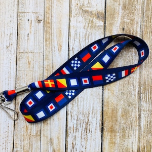 May include: A blue lanyard with a repeating pattern of nautical signal flags in red, yellow, blue, and white. The lanyard has a silver metal clip.