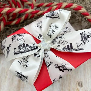 K&ouml;nnte beinhalten: Eine rote Geschenkbox mit einem wei&szlig;en Band. Das Band ist mit einem schwarz-wei&szlig;en Muster aus Mickey Mouse-Figuren, Booten und Ankern verziert.