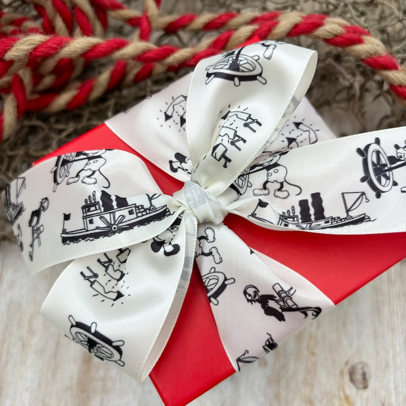 K&ouml;nnte beinhalten: Eine rote Geschenkbox mit einem wei&szlig;en Band. Das Band ist mit einem schwarz-wei&szlig;en Muster aus Mickey Mouse-Figuren, Booten und Ankern verziert.