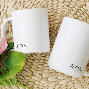 Two white ceramic mugs with black text. The mug on the left has '11 oz.' printed on the bottom. The mug on the right has '15 oz.' printed on the bottom.