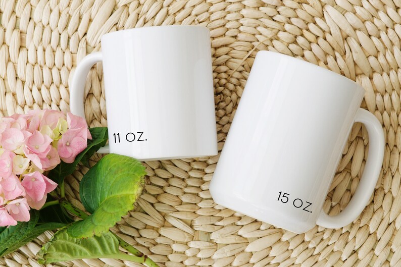 Two white ceramic mugs with black text. The mug on the left has '11 oz.' printed on the bottom. The mug on the right has '15 oz.' printed on the bottom.