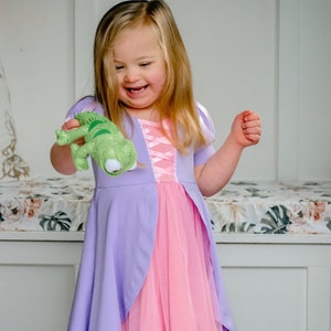 May include: A young girl wearing a purple and pink princess dress holds a green stuffed lizard toy. She is smiling and looking at the toy.