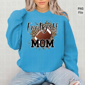 May include: A light blue sweatshirt with a graphic design of a football with a leopard print background. The text "Football Mom" is printed in black letters.