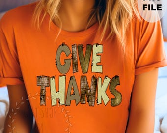 May include: An orange t-shirt featuring the phrase "GIVE THANKS" in a decorative font. The letters have a textured, plaid, and glittery design. The shirt is a solid orange colour.