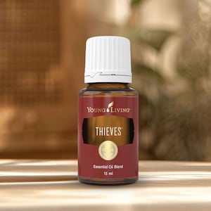 May include: A brown glass bottle of Young Living Thieves essential oil blend. The bottle has a white cap and a label with the word "THIEVES" in gold lettering. The bottle contains 15 ml of essential oil.