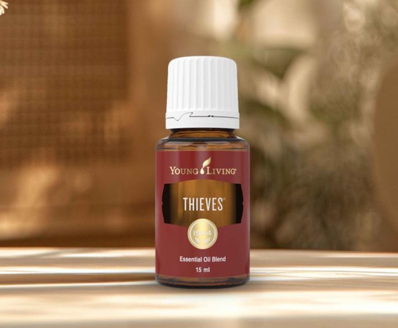 May include: A brown glass bottle of Young Living Thieves essential oil blend. The bottle has a white cap and a label with the word "THIEVES" in gold lettering. The bottle contains 15 ml of essential oil.