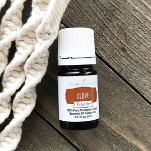 May include: A small, dark glass bottle of Young Living Clove Vitality essential oil, with a white cap and a label that reads "CLOVE Vitality." The bottle is 0.17 fl oz (5 ml). A cream-colored macrame rope is in the background.