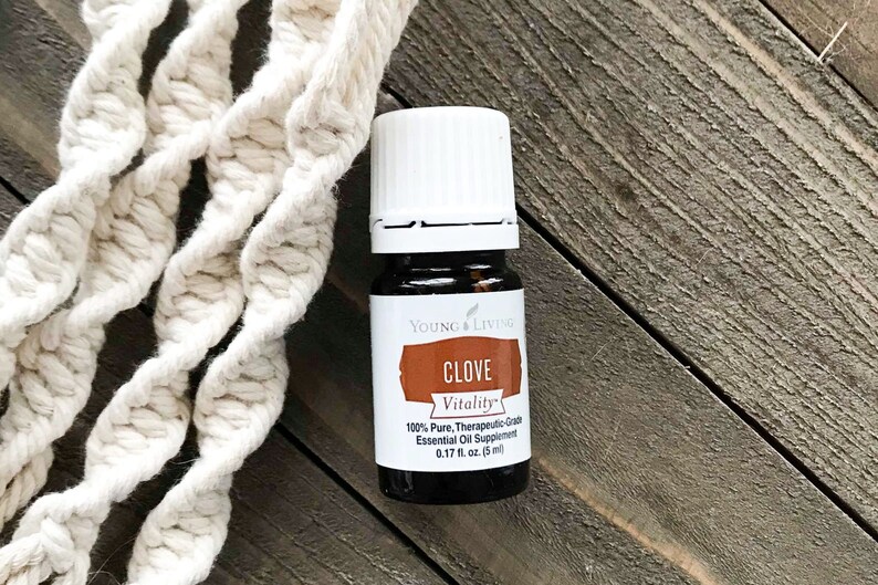 May include: A small, dark glass bottle of Young Living Clove Vitality essential oil, with a white cap and a label that reads "CLOVE Vitality." The bottle is 0.17 fl oz (5 ml). A cream-colored macrame rope is in the background.