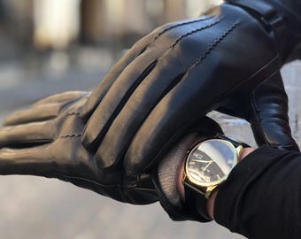 Pode incluir: Um par de luvas de couro preto com costuras decorativas no dorso da mão, usadas com um relógio de pulso dourado com mostrador preto, índices dourados e pulseira de couro preto, visível sob o punho da luva.