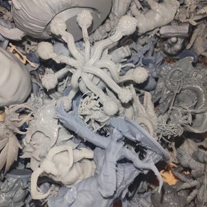 May include: A collection of unpainted, gray miniature figurines. The assortment includes fantasy creatures, such as dragons, insects, and other otherworldly beings. Some figures have intricate details, while others are more abstract.