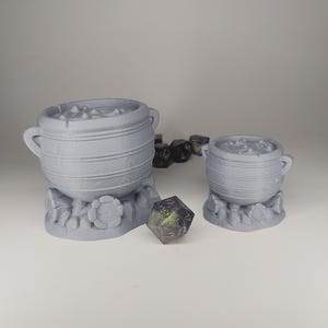 May include: Two gray, cauldron-shaped containers with handles, resting on floral bases. A multi-sided die and several black dice are next to the cauldrons. The cauldrons appear to be filled with a bubbly substance.