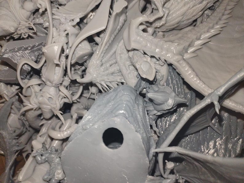 May include: A collection of unpainted, gray plastic miniatures, including dragons, fantasy creatures, and architectural elements. The pieces are intricately detailed, with visible wings, horns, and scales. The image suggests a hobby or model-making theme.