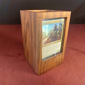 May include: A wooden card holder displaying a card with the text "Kyzaios and Tiro of Meletis." The holder is crafted from light brown wood, showcasing its grain. The card is visible through a rectangular aperture.