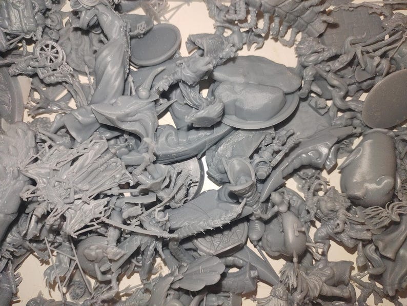 May include: A pile of unassembled gray plastic miniatures, likely for tabletop gaming. The components include figures, shields, and other detailed parts. The pieces are in various shapes and sizes, suggesting a fantasy or historical theme.