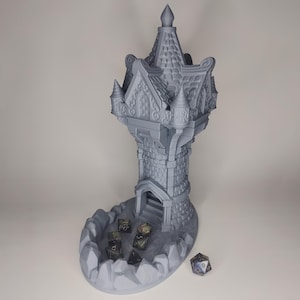May include: A grey 3D-printed dice tower, designed as a castle. The tower features a detailed stone-like texture, a crenellated roof, and a base to collect dice. Several dice are visible in the base of the tower.