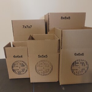May include: A collection of brown cardboard boxes of varying sizes, labeled with dimensions such as 4x4x4 inches, 5x5x5 inches, 6x6x6 inches, 7x7x7 inches, and 8x8x8 inches. Each box features a circular logo. The boxes are arranged on a dark surface.