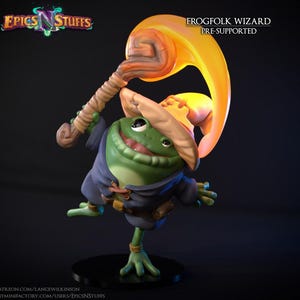 Peut inclure: Figurine de magicien grenouille verte avec un bâton et une grande flamme orange incurvée. La grenouille porte une robe bleue et un chapeau beige. Le texte "FROGFOLK WIZARD PRE-SUPPORTED" est visible.