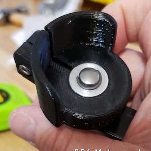 Puede incluir: Una pieza negra impresa en 3D con un botón plateado en el centro. La pieza está diseñada para ser utilizada con un coche. BRG Motorsports está impreso en la parte inferior de la pieza.