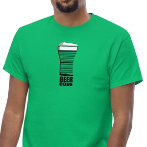 Può includere: Una maglietta verde con un disegno grafico in bianco e nero di un bicchiere da birra, progettato come un codice a barre. Il testo "BEER CODE" è stampato sotto. La maglietta è fatta di un materiale morbido.