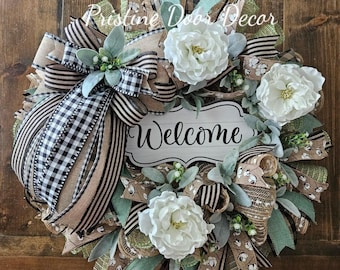 May include: A green, brown and white wreath with a burlap base and a sign that says "Welcome". The wreath is decorated with white flowers, green leaves and black and white checked ribbon.