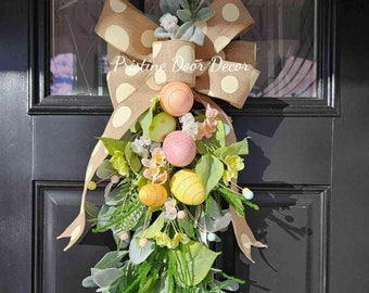 May include: A brown and white polka dot bow with a green and white floral swag and pastel colored Easter eggs. The swag is hanging on a black door. "Pristine Door Decor" is written on the bow.