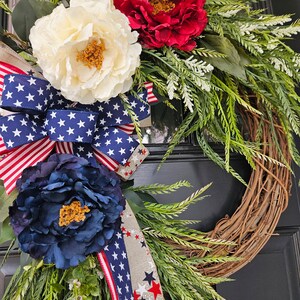 May include: A patriotic wreath with red, white, and blue flowers and a ribbon with stars. The wreath is made of grapevine and is decorated with greenery.