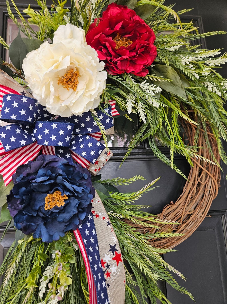 May include: A patriotic wreath with red, white, and blue flowers and a ribbon with stars. The wreath is made of grapevine and is decorated with greenery.