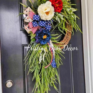 May include: A patriotic wreath with red, white, and blue flowers and a star-spangled ribbon. The wreath is hanging on a black door.