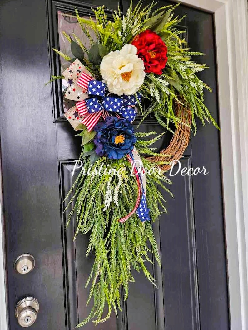 May include: A patriotic wreath with red, white, and blue flowers and a star-spangled ribbon. The wreath is hanging on a black door.