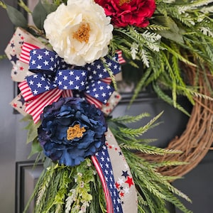 May include: A patriotic wreath with red, white, and blue flowers and a ribbon with stars. The wreath is made of twigs and has a green base.
