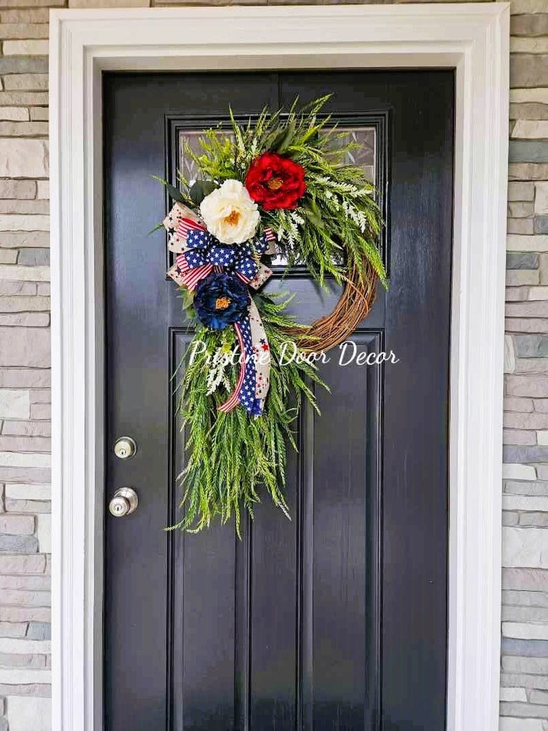 May include: A patriotic wreath with red, white, and blue flowers and a bow. The wreath is hanging on a black door with a white frame. The text "Pristine Door Decor" is visible in the bottom right corner of the image.