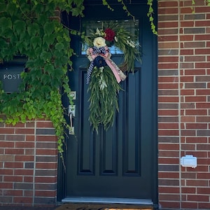 May include: A patriotic wreath with red, white, and blue ribbon and flowers hangs on a black front door. The wreath is made of greenery and has a white and blue bow. The door is surrounded by brick and ivy.