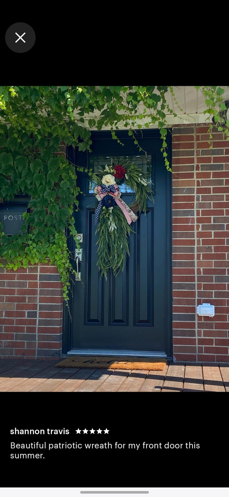 May include: A patriotic wreath with red, white, and blue ribbon and flowers hangs on a black front door. The wreath is made of greenery and has a white and blue bow. The door is surrounded by brick and ivy.