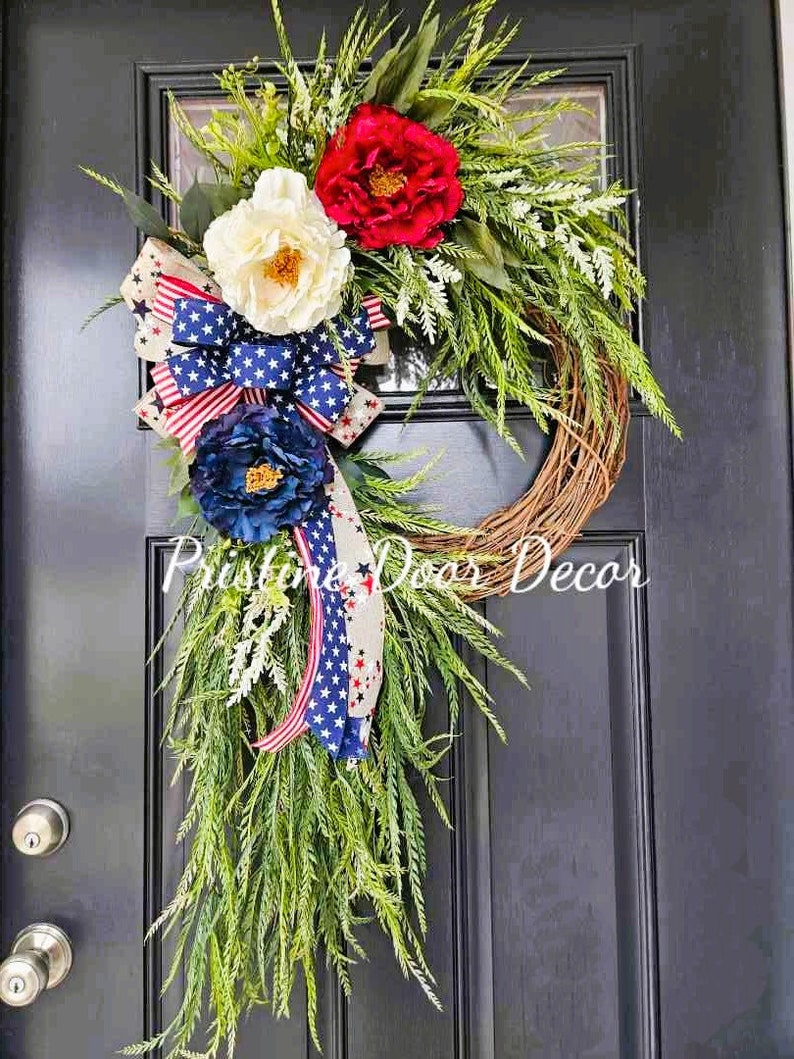 May include: A patriotic wreath with red, white, and blue flowers and a star-spangled ribbon. The wreath is made of twigs and has a green leafy base. The text "Pristine Door Decor" is visible in the bottom center of the image.