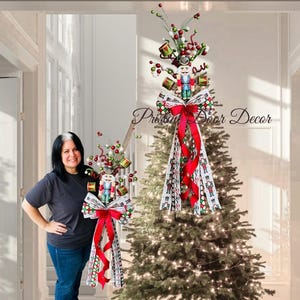 Könnte beinhalten: Weihnachtsbaumschmuck mit einem Nussknackersoldaten, einer roten Schleife und dekorativem Band. Der Schmuck hat rote, grüne und weiße Akzente, mit dem Text "Pristine Door Decor" sichtbar.