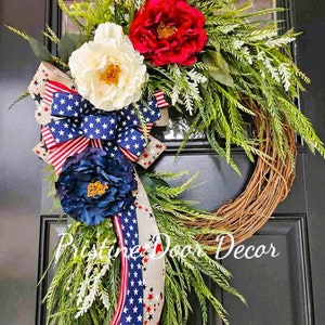May include: A patriotic wreath with red, white, and blue flowers and a ribbon with stars and stripes. The wreath is made of grapevine and is decorated with greenery. The text "Pristine Door Decor" is visible in the bottom right corner of the image.
